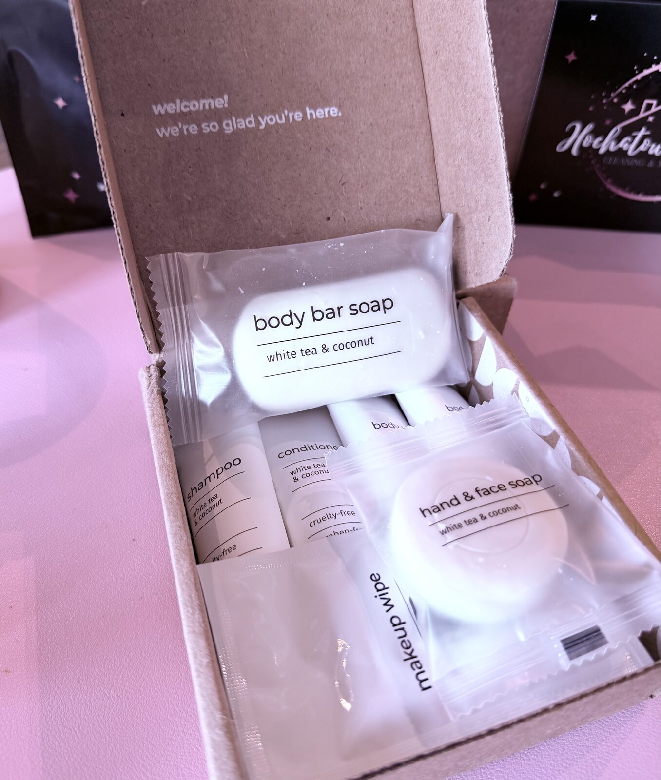 An open half-bath essentials box; the lid reads welcome, we're so glad you're here, with a body bar, shampoo, conditioner, hand and face soap and a makeup wipe inside