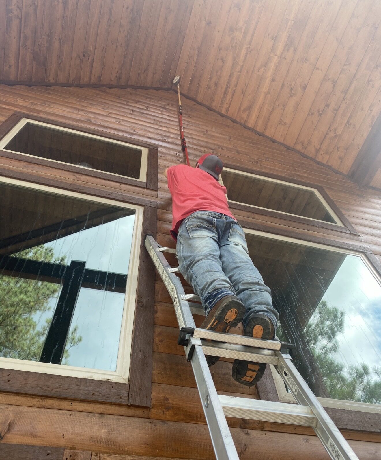 Hochatown Premier crew member on an extension ladder using a water-fed pole to wash the high windows of a wood cabin