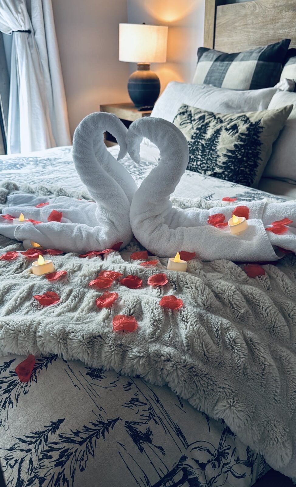 Two white towels folded into swans forming a heart, with red rose petals and flameless tea lights on a plush throw