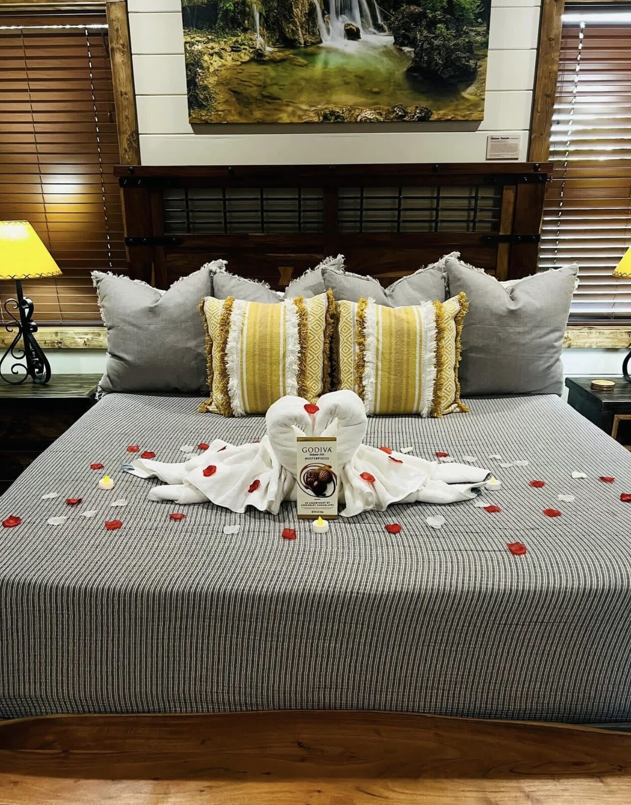 King bed styled with towel swans, a Godiva chocolate box and scattered rose petals beneath a large framed waterfall photograph